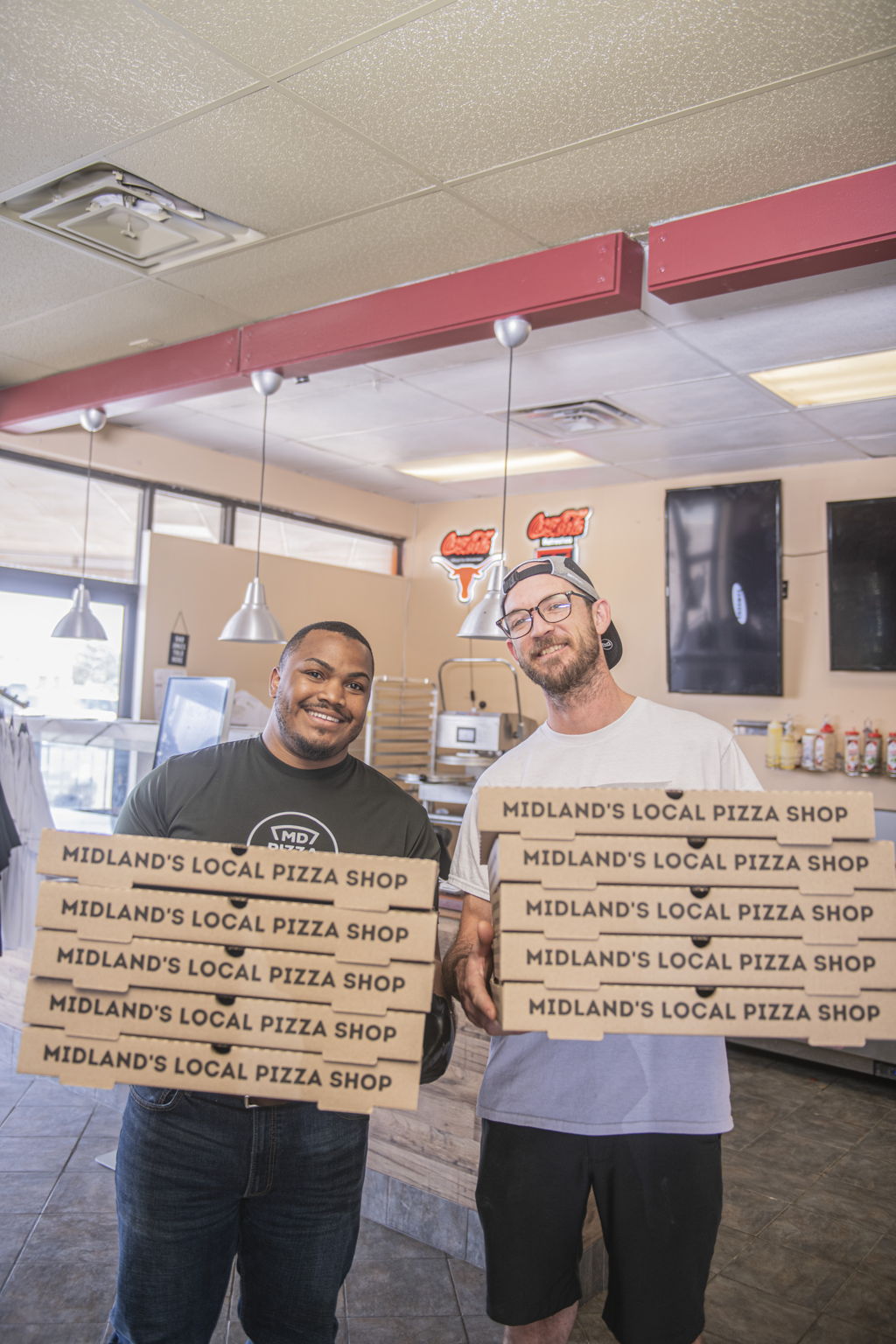 Ruston and Dru in the Midland pizza shop, each holding a stack of Midland's Local Pizza Shop boxes